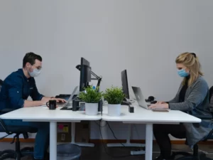 Employees working face to face with laptops and mask on in corporate office